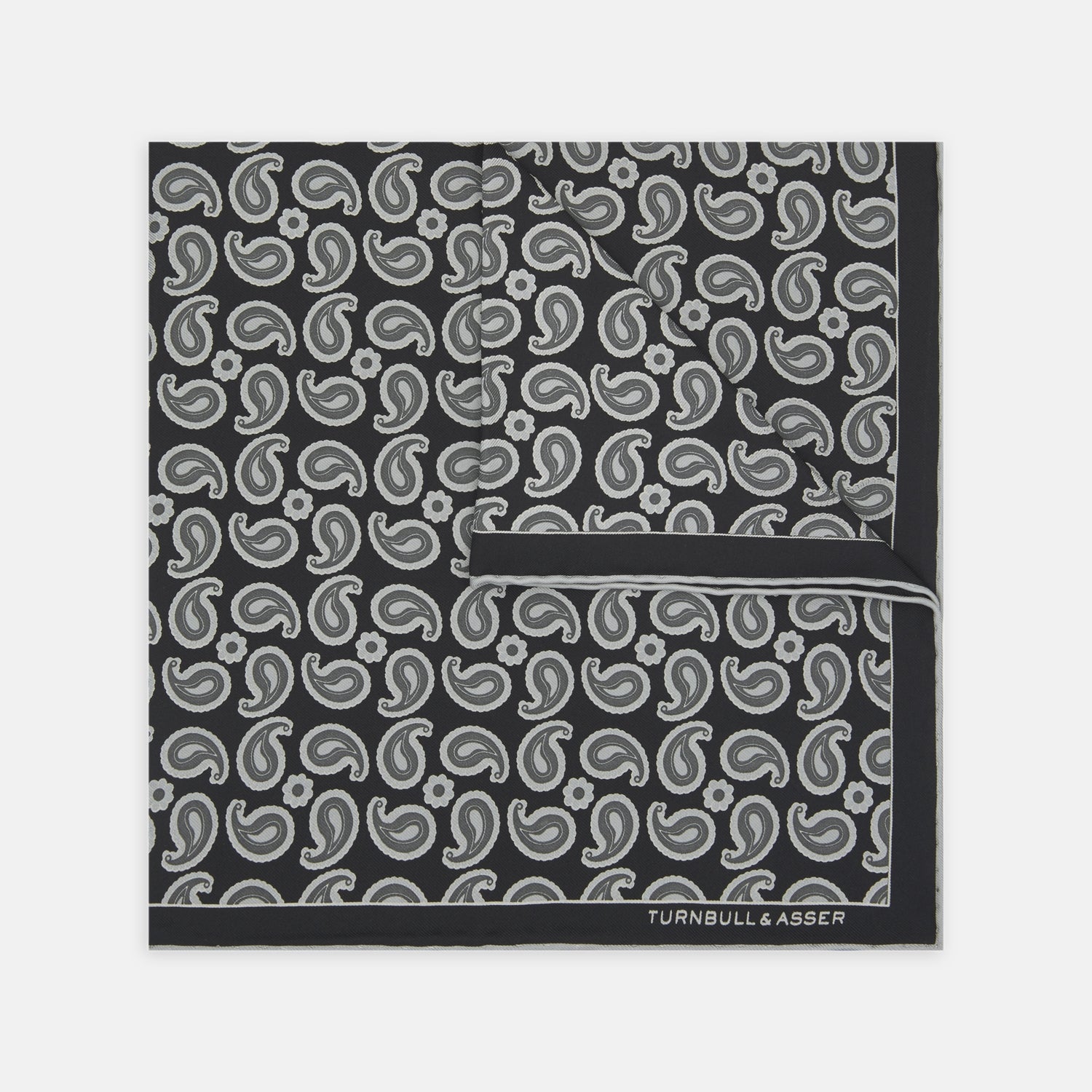 View 2 of product The Monochrome Paisley Mill Silk Pocket Square by Turnbull & Asser is a limited edition black silk accessory with a white paisley and floral pattern. The brand name appears in white text on the slightly folded bottom right corner.