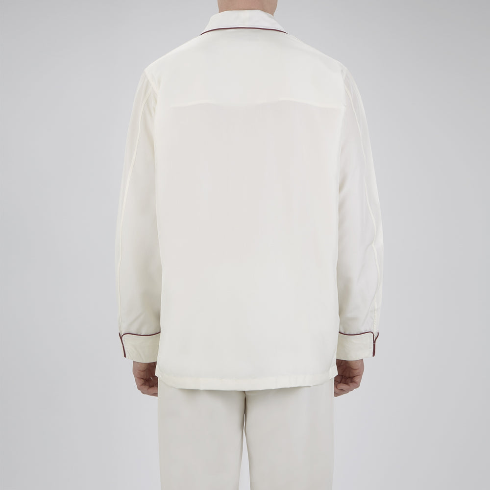 The Turnbull & Asser Cream Piped Silk Pyjama Set is neatly folded with dark piping and buttons, placed next to a matching white sleep mask with tie straps, all set against a light neutral background.