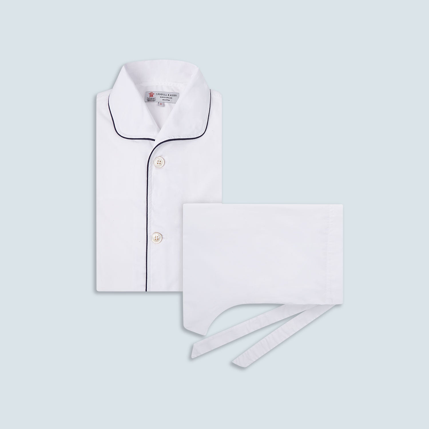 View 1 of product The Turnbull & Asser White Piped Cotton Pyjama Set, made from Egyptian cotton and featuring black piping and two buttons, is shown folded with a matching white tie-strap face mask on a light gray background.