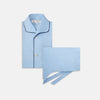 View 1 of product The Light Blue Piped Cotton Pyjama Set by Turnbull & Asser is neatly folded, with dark piping on the collar and buttons, displayed next to a matching light blue sleep mask with tie straps on a plain white background.