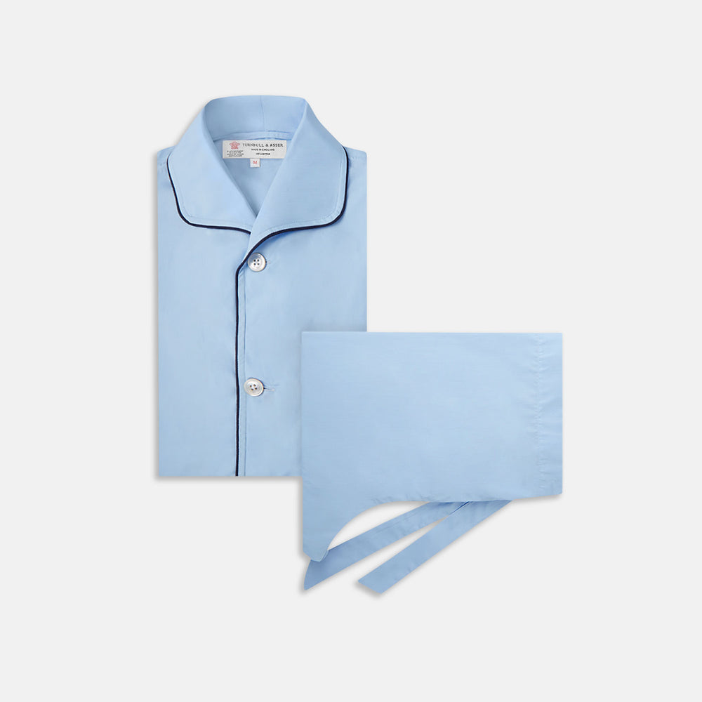 The Light Blue Piped Cotton Pyjama Set by Turnbull & Asser is neatly folded, with dark piping on the collar and buttons, displayed next to a matching light blue sleep mask with tie straps on a plain white background.