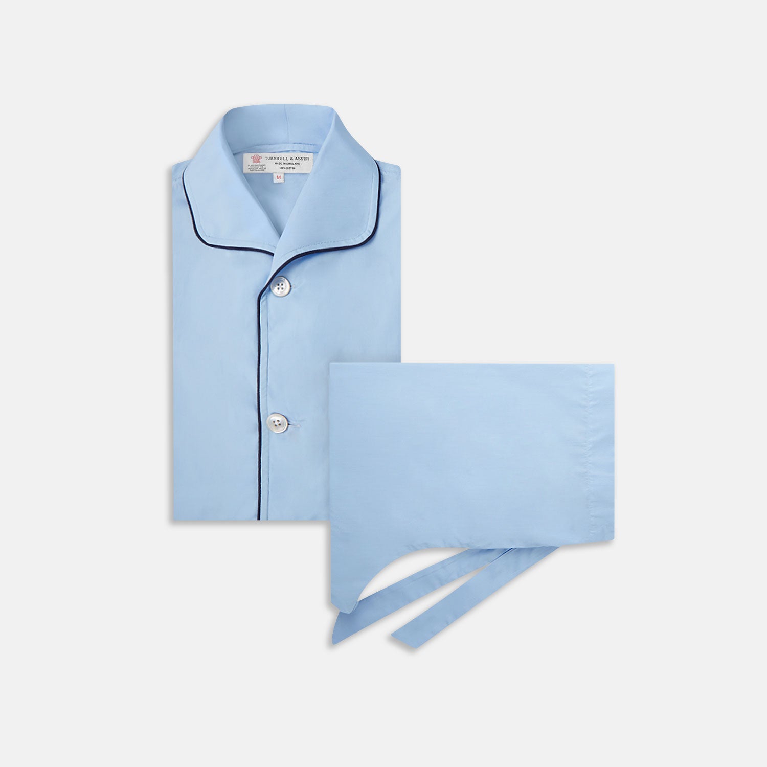 View 1 of product The Light Blue Piped Cotton Pyjama Set by Turnbull & Asser is neatly folded, with dark piping on the collar and buttons, displayed next to a matching light blue sleep mask with tie straps on a plain white background.