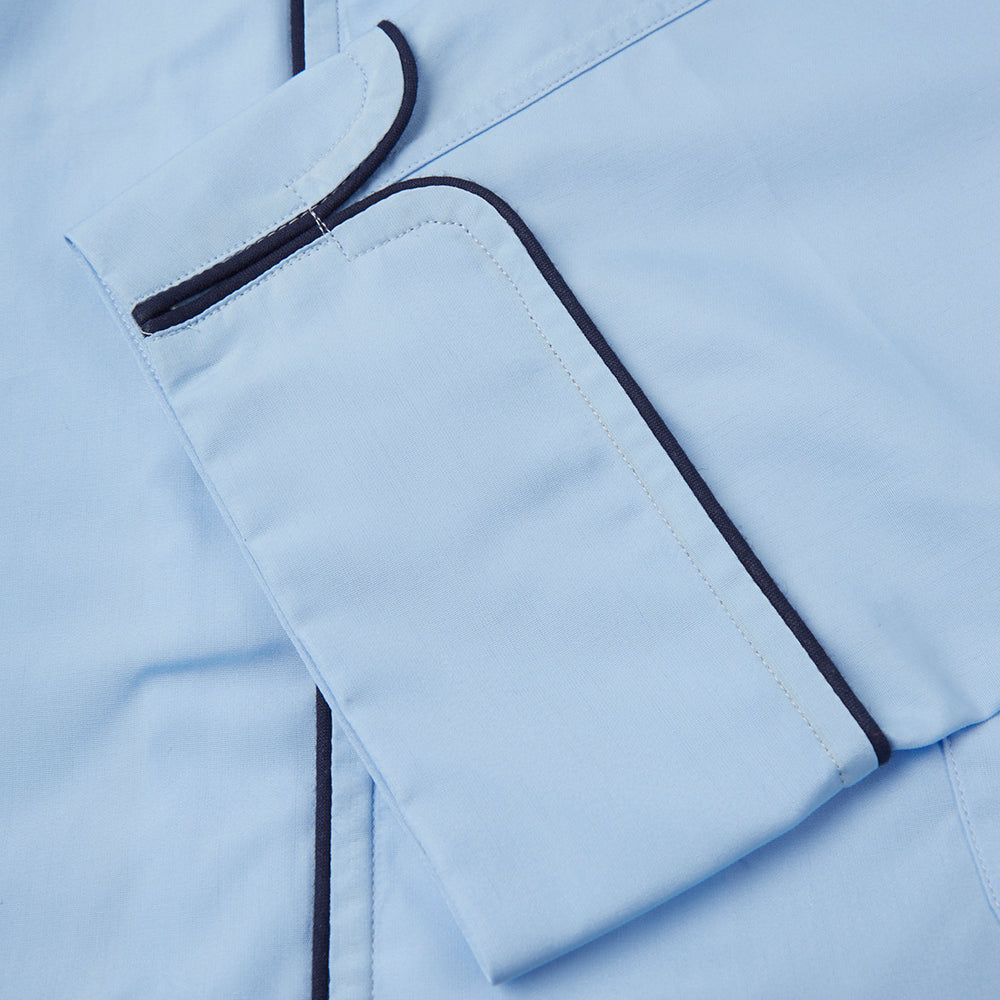 The Light Blue Piped Cotton Pyjama Set by Turnbull & Asser is neatly folded, with dark piping on the collar and buttons, displayed next to a matching light blue sleep mask with tie straps on a plain white background.