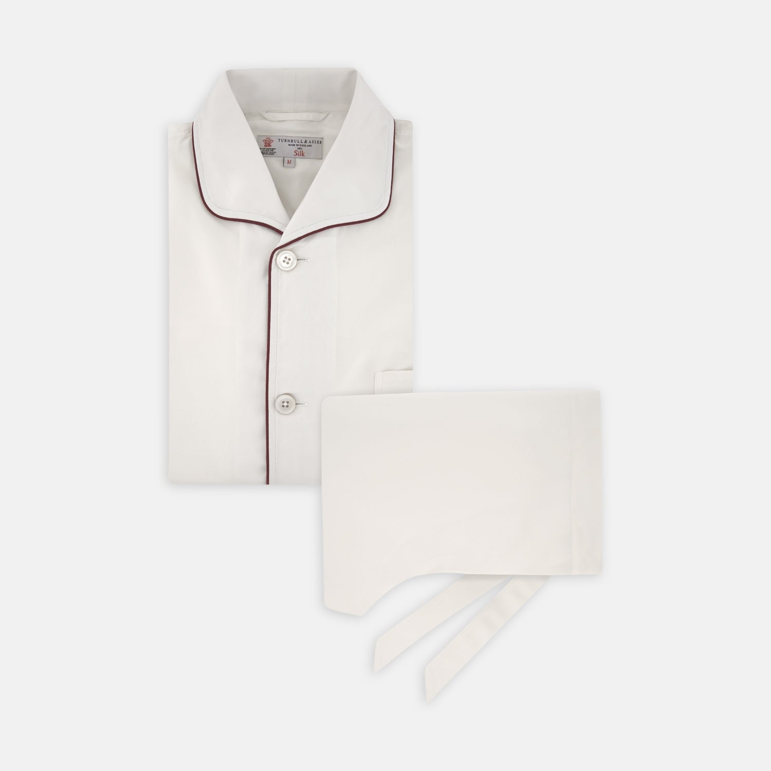 View 1 of product The Turnbull & Asser Cream Piped Silk Pyjama Set is neatly folded with dark piping and buttons, placed next to a matching white sleep mask with tie straps, all set against a light neutral background.