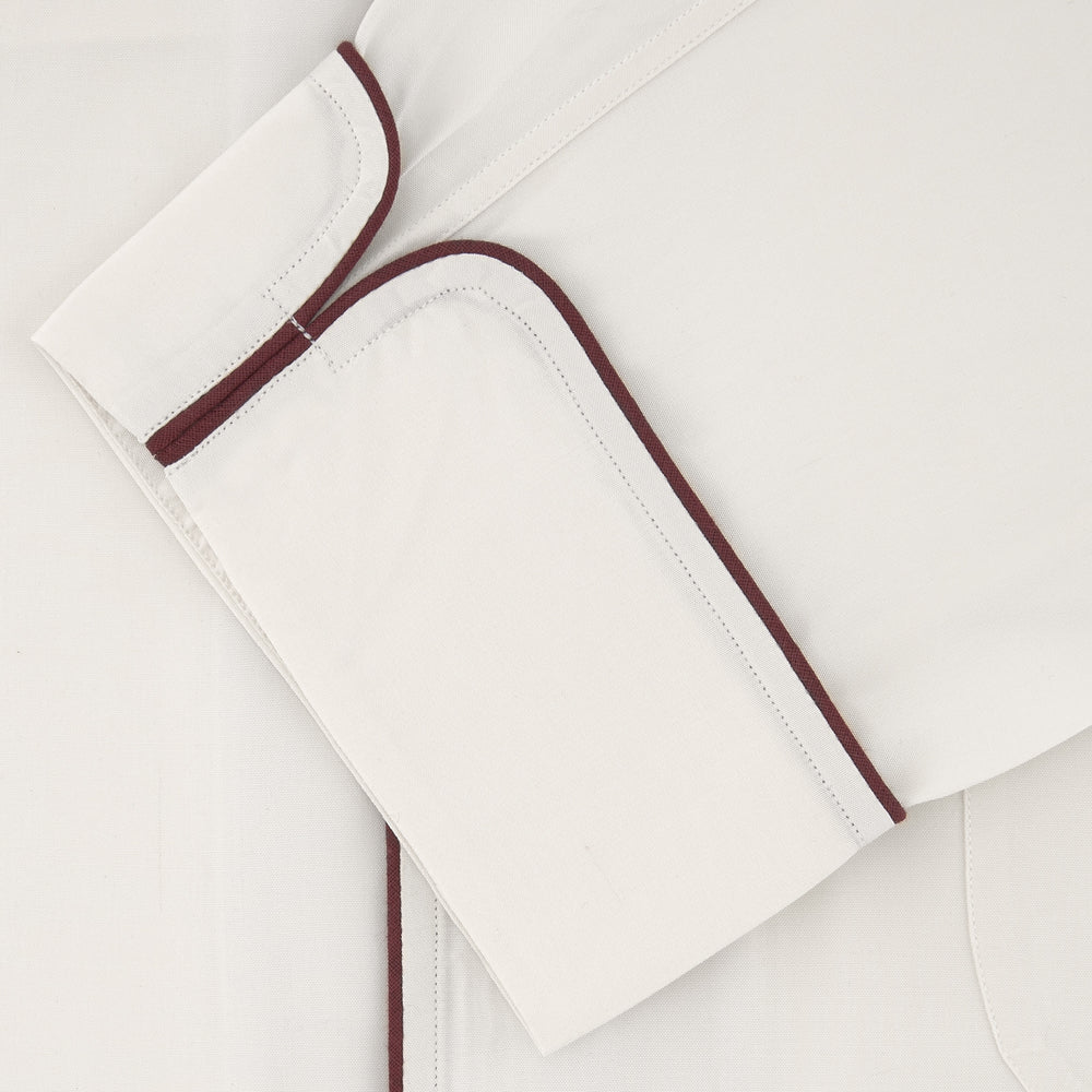 The Turnbull & Asser Cream Piped Silk Pyjama Set is neatly folded with dark piping and buttons, placed next to a matching white sleep mask with tie straps, all set against a light neutral background.