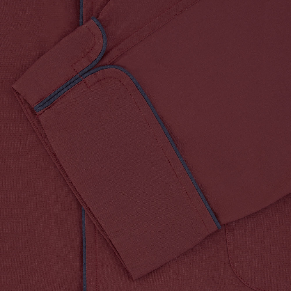 The Wine Piped Silk Pyjama Set by Turnbull & Asser features a folded maroon top with white buttons and dark trim, displayed beside a matching sleep mask with tie straps, set against a plain white background.