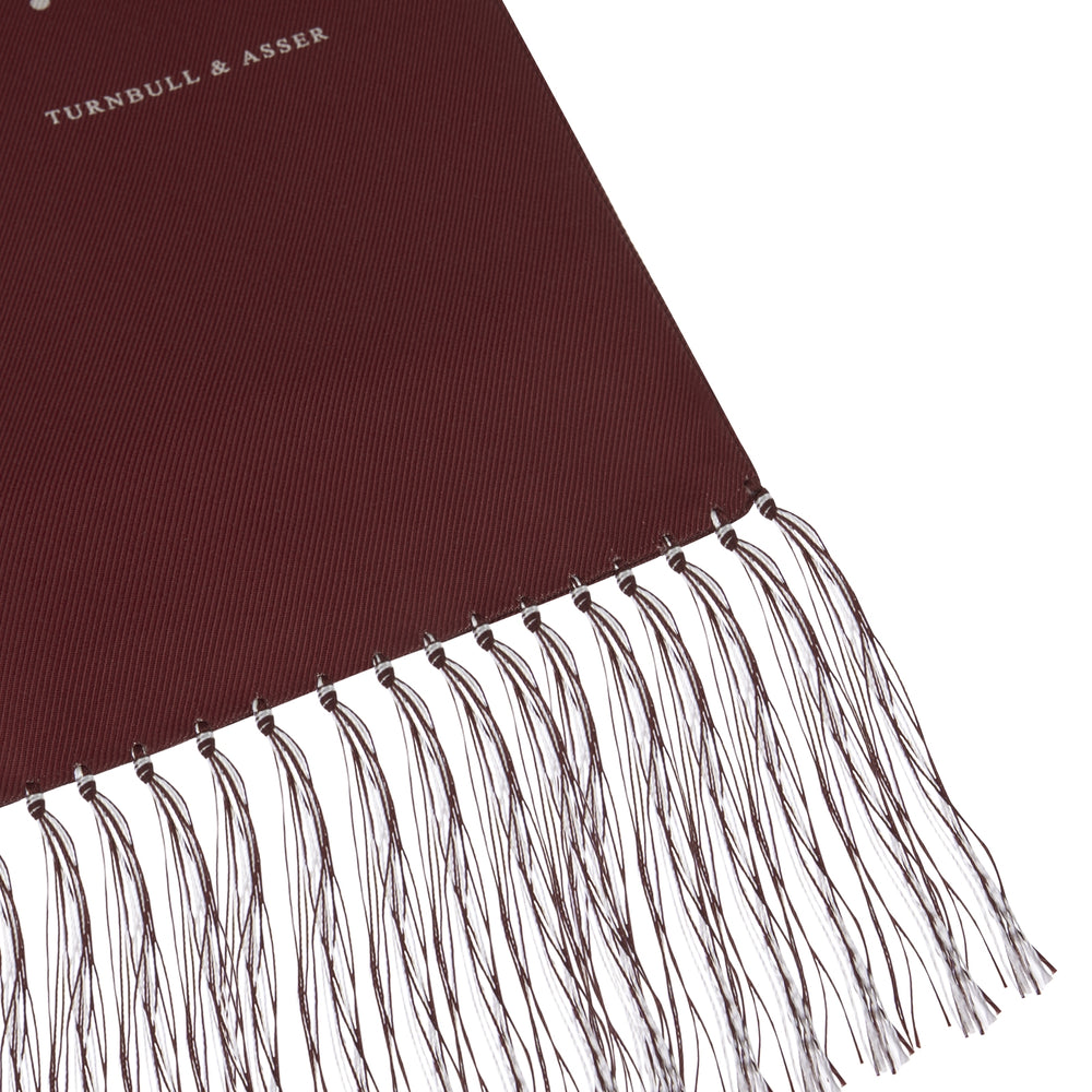 The Turnbull & Asser Wine Spotted Silk Scarf features white polka dots and white fringed ends, elegantly tasseled and neatly folded against a plain white background.