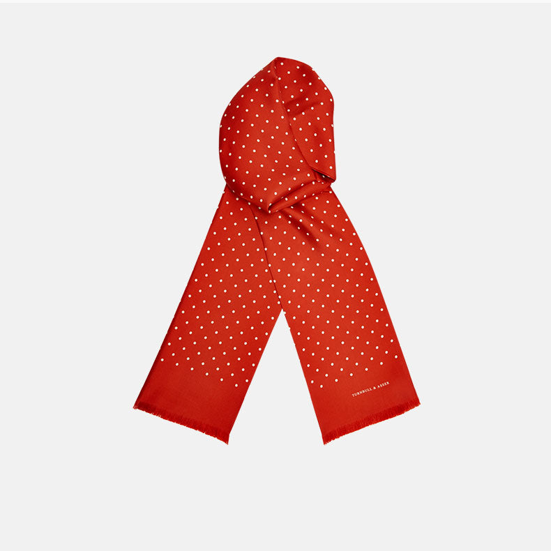 View 1 of product The Red and White Spotted Silk Scarf by Turnbull & Asser features white polka dots and fringed edges on silk. The brand name appears subtly on one end, with the scarf displayed against a plain light background.