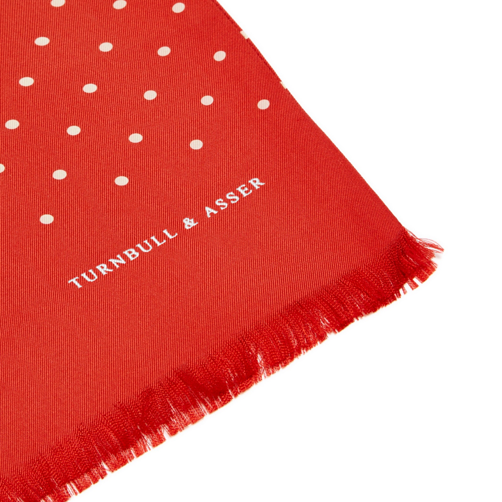 View 2 of product The Red and White Spotted Silk Scarf by Turnbull & Asser features white polka dots and fringed edges on silk. The brand name appears subtly on one end, with the scarf displayed against a plain light background.