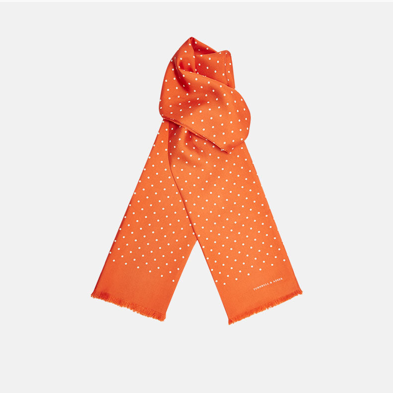 View 1 of product The Turnbull & Asser Orange and White Spotted Silk Scarf, featuring white polka dots and fringed edges, is neatly folded and displayed on a plain light gray background.
