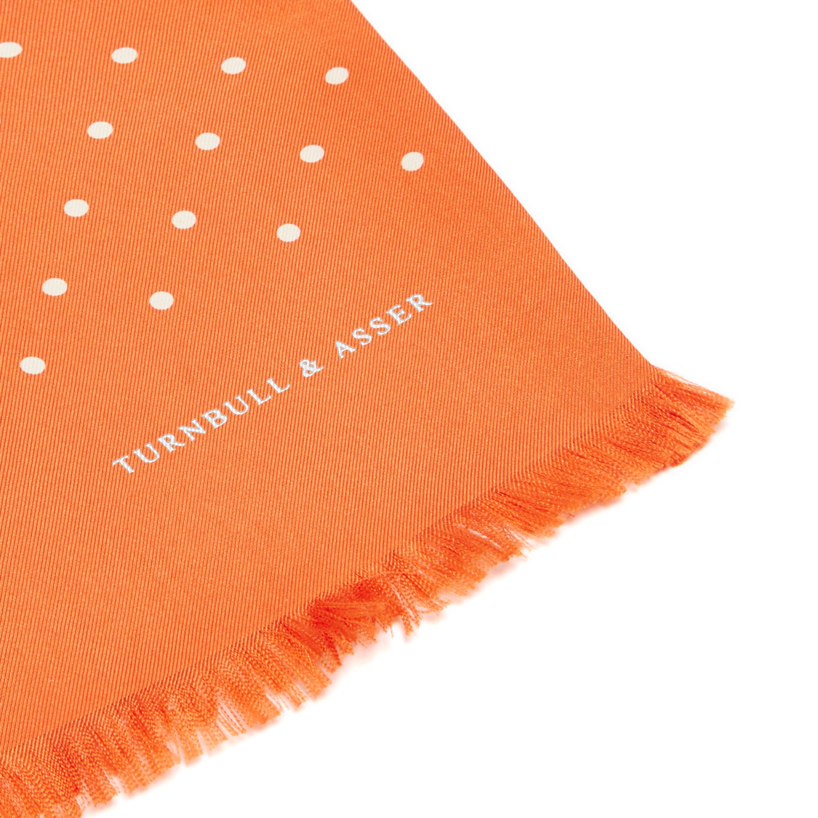 View 2 of product The Turnbull & Asser Orange and White Spotted Silk Scarf, featuring white polka dots and fringed edges, is neatly folded and displayed on a plain light gray background.