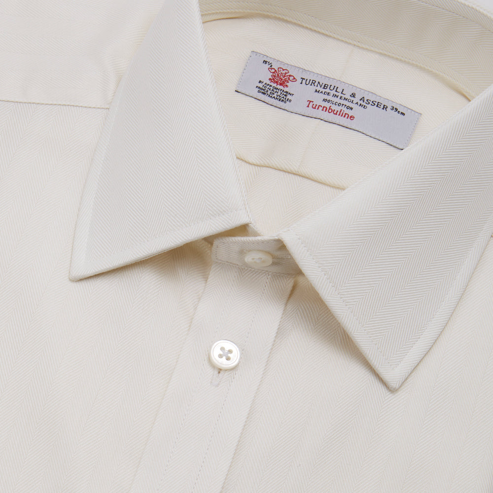 The Turnbull & Asser Cream Herringbone Superfine Cotton Shirt features a T&A collar, double cuffs, and a subtle pattern. It is neatly folded with white buttons and displayed on a plain white background.