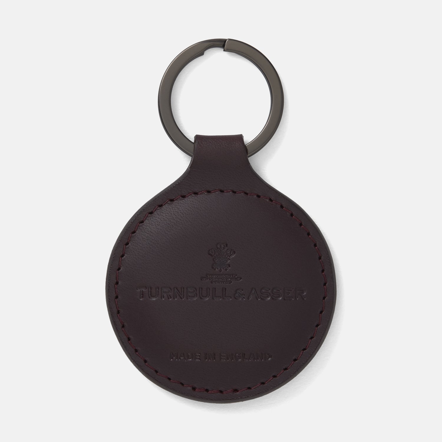 View 1 of product The Violla Leather Key Fob by Turnbull & Asser is a round, dark brown leather accessory with embossed branding, visible edge stitching, and a metal loop at the top. Made in England, it combines elegance and function for your keys.