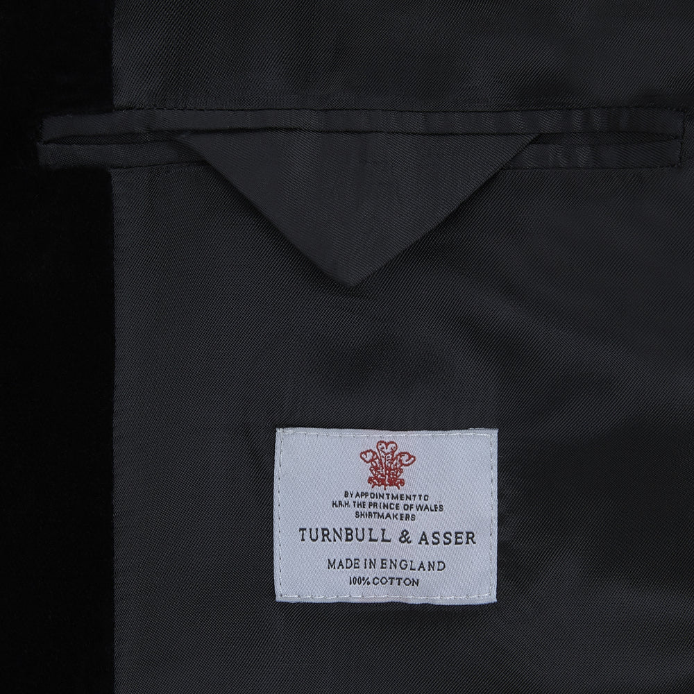 The Turnbull & Asser Black Single Breasted Velvet Smoking Jacket features a shawl collar, satin lining, two front pockets, and a knotted closure—ideal for stylish eveningwear.