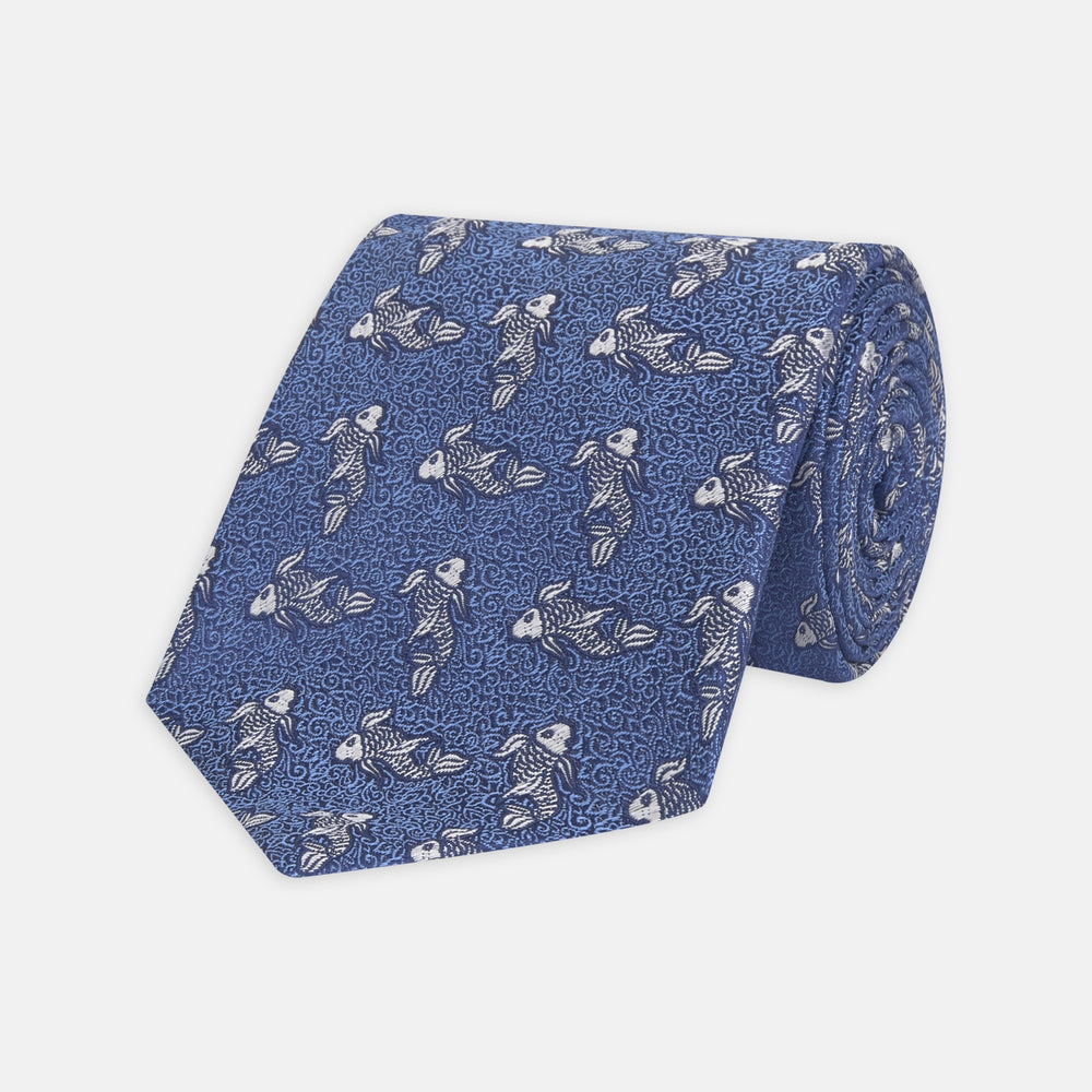 The Blue Carp Silk Tie by Turnbull & Asser features a blue silk base with white and dark blue koi fish among light blue swirls. The tie is displayed rolled up, pointed end forward.