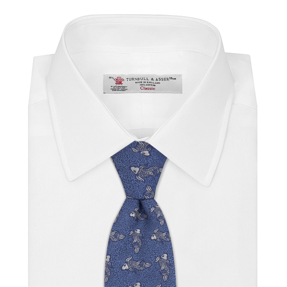 The Blue Carp Silk Tie by Turnbull & Asser features a blue silk base with white and dark blue koi fish among light blue swirls. The tie is displayed rolled up, pointed end forward.