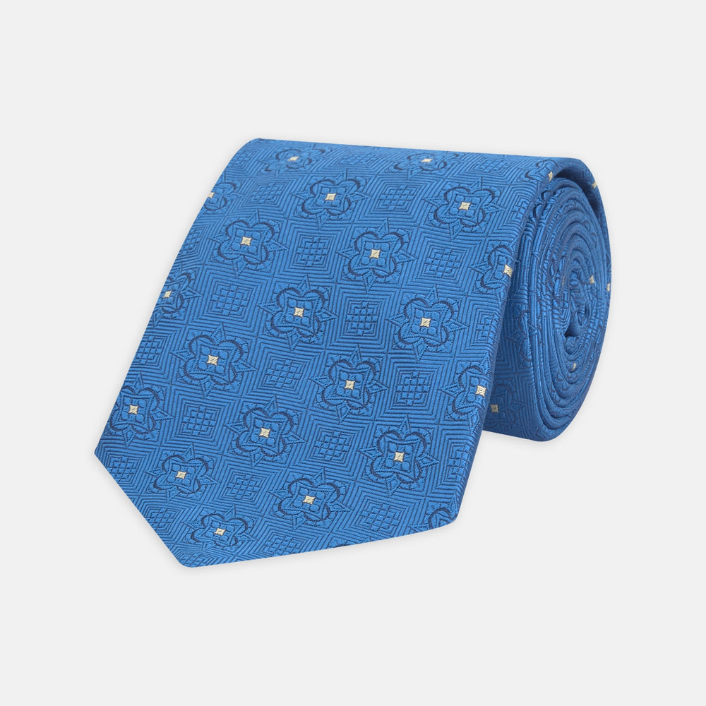 The Turnbull & Asser Blue Flower Mosaic Silk Tie features a blue silk fabric with a detailed geometric and floral design, accented in lighter blue and white, displayed on a white background.